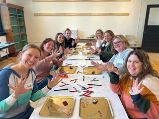group of women at jewelry workshop sitting at a table with trays that contain items needed to make a necklace and earring set
